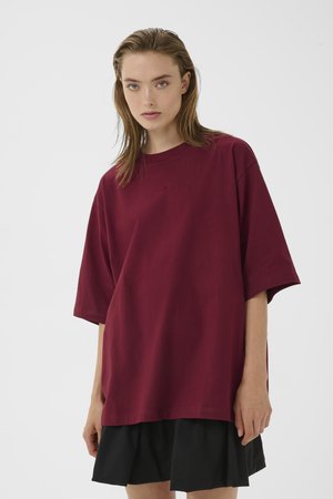 Jeune femme aux cheveux mouillés portant un t-shirt bordeaux oversize et une jupe plissée noire, debout devant un fond blanc uni.
