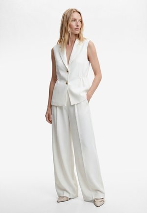 Donna che indossa un blazer bianco senza maniche con due bottoni e pantaloni bianchi a gamba larga, in piedi con una mano in tasca.