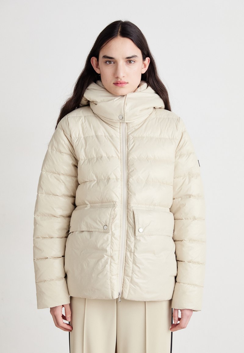 Casaco puffer bege com colarinho alto, fecho de correr na frente e bolsos com botão de pressão. Apresenta textura acolchoada horizontal e acabamento liso e brilhante.