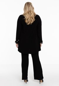 Zwarte lange mouw tuniek met een losse pasvorm en ronde zoom, gecombineerd met flared zwarte broek. Bevat zwarte accenten op de manchetten, gladde textuur.