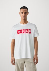 T-shirt en coton blanc avec un logo rouge "DIESEL" et le texte "DENIM DIVISION". Manches courtes, encolure ras du cou, coupe décontractée.