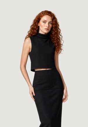 Giovane donna con lunghi capelli ricci rossi che indossa un top nero smanicato e una gonna nera a vita alta su uno sfondo grigio chiaro.