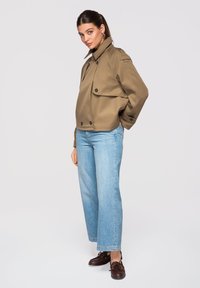 Femme portant une veste marron boutonnée, un jean bleu clair et des chaussures marron foncé, debout devant un fond uni.