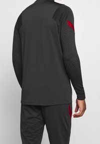 Langærmet sports top i mørkegrå med teksturerede sorte detaljer på skuldrene og røde sidelæg. Blødt, fleksibelt stof designet til bevægelse.