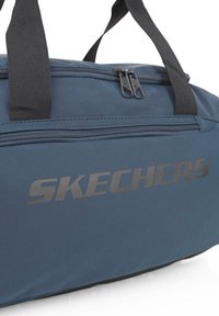 Bolsa de viaje Skechers en tela azul oscuro, con superficie suave, dobles cremalleras y un prominente logo negro en el lateral. Asas de transporte resistentes.