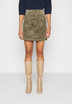 Kobieta ubrana w mini spódnicę ze sztruksu w kolorze oliwkowym, granatową bluzkę z długim rękawem oraz beżowe kozaki za kolano na obcasie, stojąca na szarej podłodze.