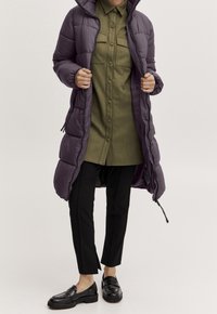 Osoba ubrana w długi fioletowy pikowany płaszcz na oliwkowej, zapinanej na guziki sukience koszulowej, czarne spodnie o długości do kostek i czarne mokasyny na neutralnym tle.