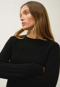 Pull noir à côtes avec des manches longues et un col rond. Texture douce avec des rayures verticales. Bras croisés, design simple et classique.