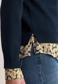 Mörkblå stickad tröja med sidoflikar i lager över en blommönstrad krämfärgad och blå skjorta, som kombineras med blå denimjeans.