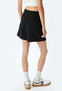 Zwarte hoog getailleerde shorts met een lichte flare, voorzien van twee achterzakken en een gladde textuur. Gecombineerd met witte sneakers en geribbelde sokken.