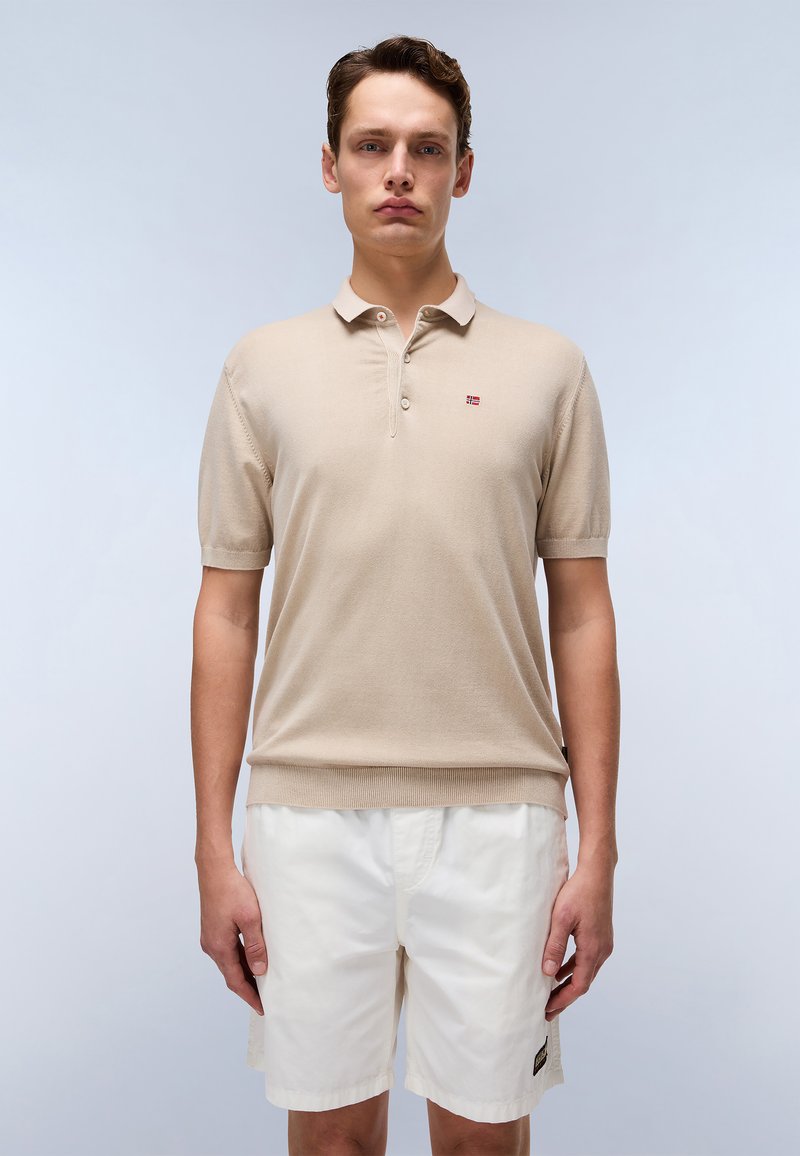 Polo shirt beige a maniche corte con colletto e tre bottoni. Tessuto a maglia dalla consistenza fine. Abbinato a pantaloni corti bianchi.