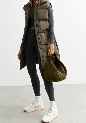 Femme portant une longue doudoune sans manches vert olive, des leggings foncés, des baskets blanches, tenant un sac matelassé assorti vert olive.