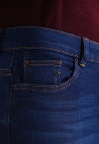 Pantalones vaqueros de mezclilla azul oscuro con un bolsillo trasero visible con costuras naranjas y un acabado texturizado, parcialmente cubiertos por una tela color granate.