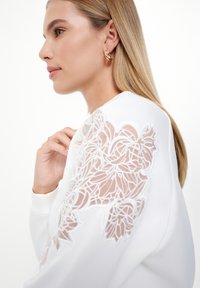 Top blanco con detalles bordados transparentes en las mangas, que presentan patrones florales. La modelo tiene el cabello largo y lleva un pendiente de aro dorado.