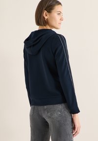 Marineblauer Hoodie mit Rückansicht, ausgestattet mit einer Kapuze mit Kordelzug, seitlichen Streifen und gerippten Bündchen. Kombiniert mit grauen Jeansstoffhosen.