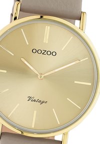 Montre dorée avec un cadran rond, un visage texturé en or clair, des repères horaires minimalistes et un bracelet en cuir marron clair. Présente les inscriptions "OOZOO" et "Vintage".