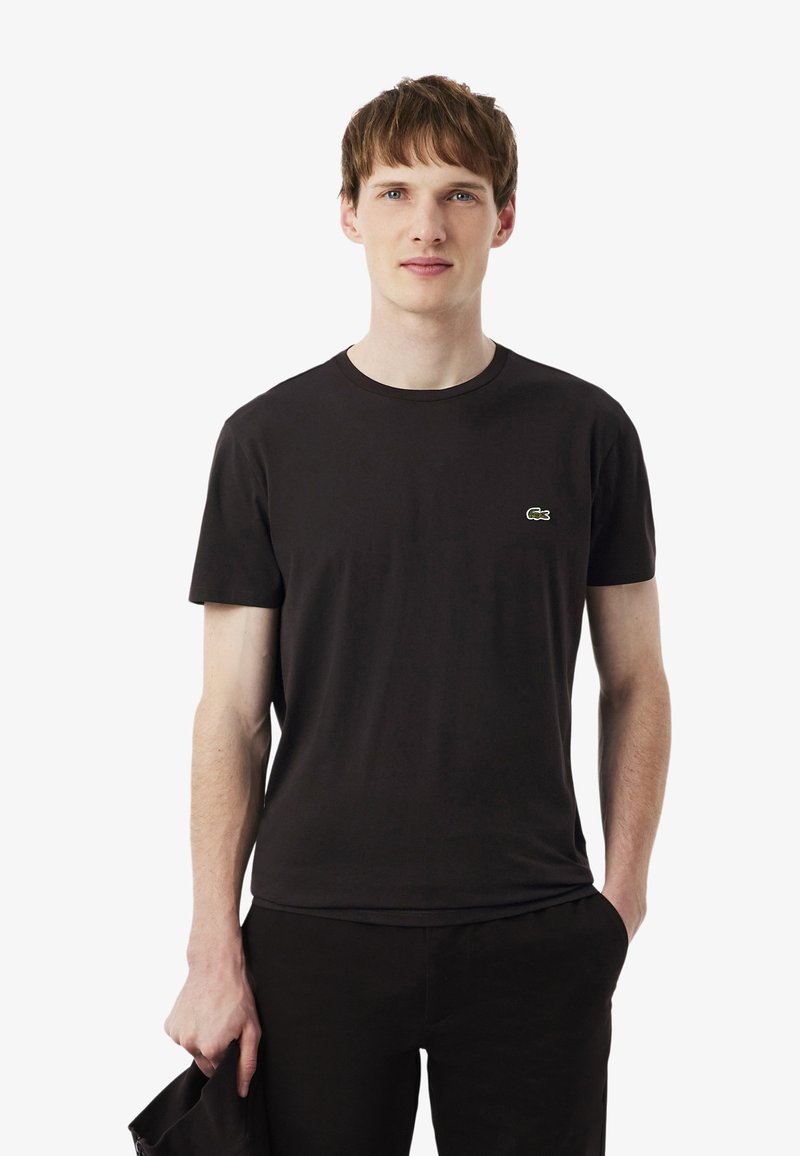 T-shirt en coton noir avec un col rond, des manches courtes et le logo vert Lacoste sur le côté gauche de la poitrine. Le modèle tient un objet noir.