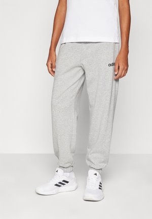 Personne portant un pantalon de survêtement Adidas gris clair et des baskets blanches Adidas, debout devant un fond clair et uni.
