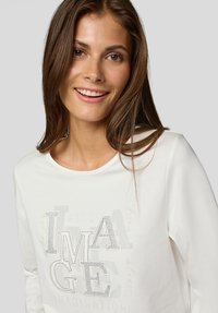 Lächelnde Frau mit langen braunen Haaren, die ein weißes Hemd mit mit Strass besetzten Buchstaben trägt, die "IMAGE" auf einem hellgrauen Hintergrund buchstabieren.