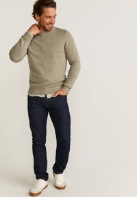Un pull en maille vert clair avec un col et des poignets côtelés, associé à un jean bleu foncé et des baskets blanches, se tenant dans une pose détendue.