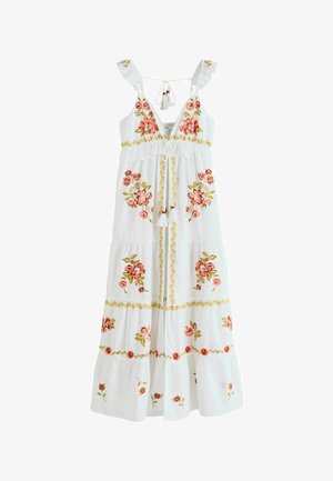 Femme debout pieds nus dans une longue robe blanche avec broderies florales, dans une porte en pierre encadrée de poutres en bois rustiques.