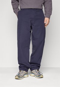 Homme portant un pantalon large bleu marine, des chaussures de trail grises et beiges, et un sweat-shirt gris, se tenant debout les mains dans les poches.