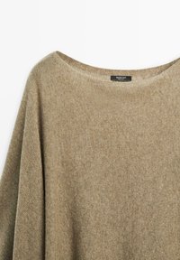 Pull en tricot beige, surdimensionné, avec un col large, fabriqué dans un matériau doux. Étiquette visible au niveau du cou, mettant en valeur une texture subtile.