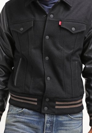 Veste noire boutonnée avec manches en cuir, poches poitrine, ceinture côtelée rayée, portée avec un jean bleu.