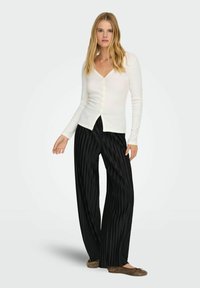 Kobieta w białym kardiganie zapinanym na guziki, czarnych plisowanych spodniach o szerokich nogawkach oraz brązowych balerinach, stojąca na jednolitym tle.