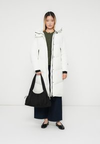 Kvinde i lang hvid polstret frakke, iført sort håndtaske, mørke brede bukser og sorte balletflade sko, stående foran en hvid baggrund.