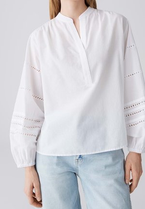 Kvinde iført hvid bluse med lange ærmer og hulmønster på ærmerne samt lyseblå jeans, vist fra skuldre til hofter.