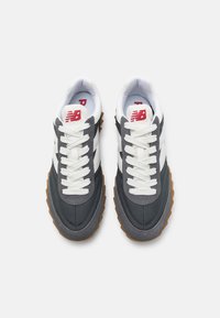 Zapatos deportivos grises y blancos con una parte superior texturizada, suela de goma y acentos en beige contrastantes. Presentan cordones y detalle del logo en la lengua.