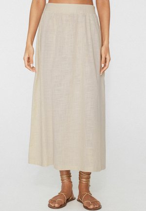 Falda midi de lino beige combinada con sandalias planas doradas con tiras, mostrada desde la cintura hasta los pies sobre un fondo neutro.