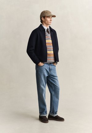 Mann mit beiger Kappe, gemusterter Strickweste über Hemd und Krawatte, schwarzer Jacke, blauen Jeans und dunkelbraunen Schuhen, stehend mit Händen in den Taschen.