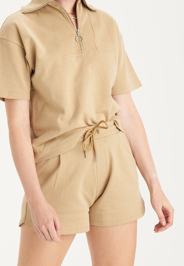 Béžový bavlněný outfit obsahující tričko s krátkým rukávem a půlzipovým límcem, spolu s odpovídajícími šortkami na šňůrku v uvolněném střihu.