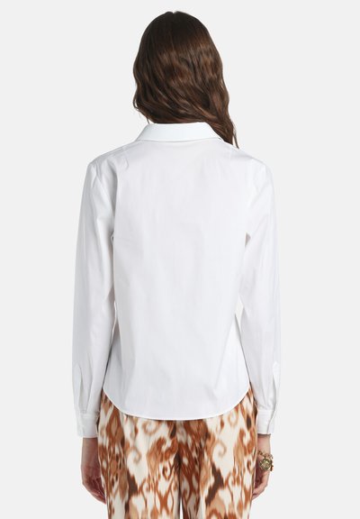 White long-sleeve shirt with a collared neck, featuring a curved hem and buttoned cuffs, paired with brown patterned pants.