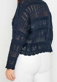 Marineblauer, gestrickter Cardigan mit einem offenen Häkelmuster, langen Ärmeln und gewelltem Saum. Über weißer Kleidung getragen für einen Kontrast.
