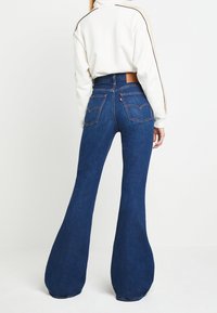 Rozšířené modré džíny s vysokým pasem, výrazným prošíváním a dvěma zadními kapsami. Tkanina vypadá texturovaně a má střední hmotnost.