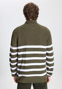 Hombre con cabello castaño rizado, vistiendo un suéter verde oliva con rayas horizontales blancas, mostrado desde la espalda contra un fondo liso.