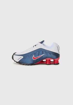 Botte de sport Nike avec un dessus blanc agrémenté d'accents bleu marine et rouge, texture perforée et une semelle rouge striée distinctive.