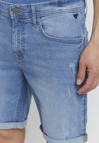 Denimshorts i lys blå med slitt finish, rulleknotter og en liten svart logo nær lommen. Har klassisk femlommers design.