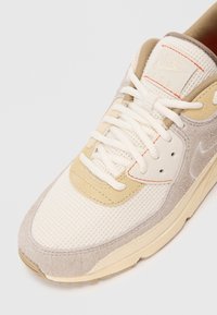 Gros plan d'une basket Nike Air beige et blanche avec des détails en mesh et en daim, des lacets blancs et des coutures orange sur la languette.
