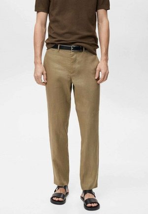 Mann trägt hellbraune Hose, dunkelbraunes kurzärmeliges Hemd, schwarzen Gürtel und schwarze offene Sandalen, steht vor weißem Hintergrund.