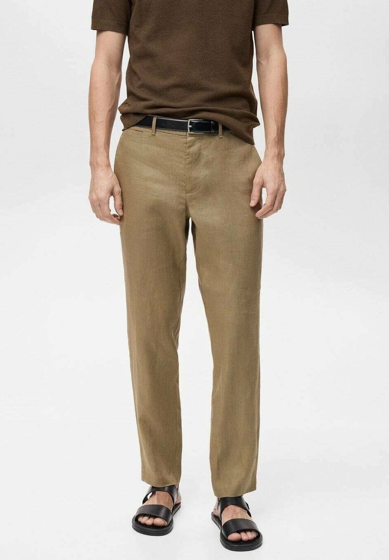 Homme portant un pantalon marron clair, une chemise à manches courtes marron foncé, une ceinture noire et des sandales noires à bouts ouverts, debout sur fond blanc.
