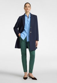 Abrigo navy, camisa de rayas azul claro, pantalones verdes de corte ajustado y tacones marrones. El abrigo tiene cierre de botones; la camisa cuenta con detalle en el cuello y un bolsillo.