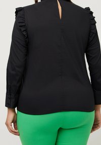 Blouse noire à manches longues avec des accents froncés aux épaules, ouverture en forme de clé au dos, accompagnée d'un pantalon moulant vert vif. Tissu lisse et léger.