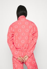 Korallfärgad vindjacka med elastiska ärmslut och midja, prydd med vita cirkulära logotyper över hela. Visad bakifrån.