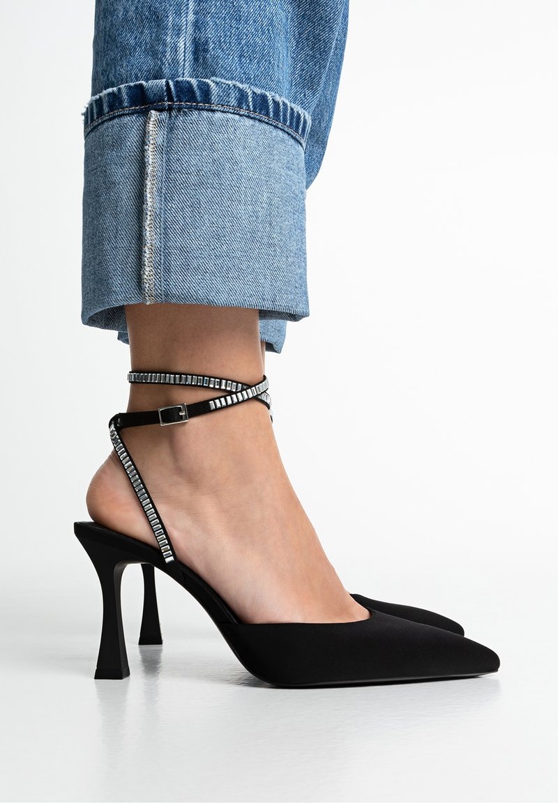 Pied portant un escarpin noir à bout pointu avec une bride de cheville cloutée carrée, associé à un jean en denim bleu retroussé.