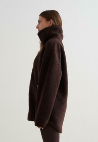 Bruine fleecetrui met een hoge kraag, overlengte ontwerp, textuur oppervlak en zijrits detail, weergegeven tegen een lichte muur als achtergrond.
