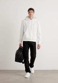 Felpa bianca in cotone con tasca a marsupio, pantaloni neri e sneaker bianchi. Il modello porta uno zaino in pelle nera.